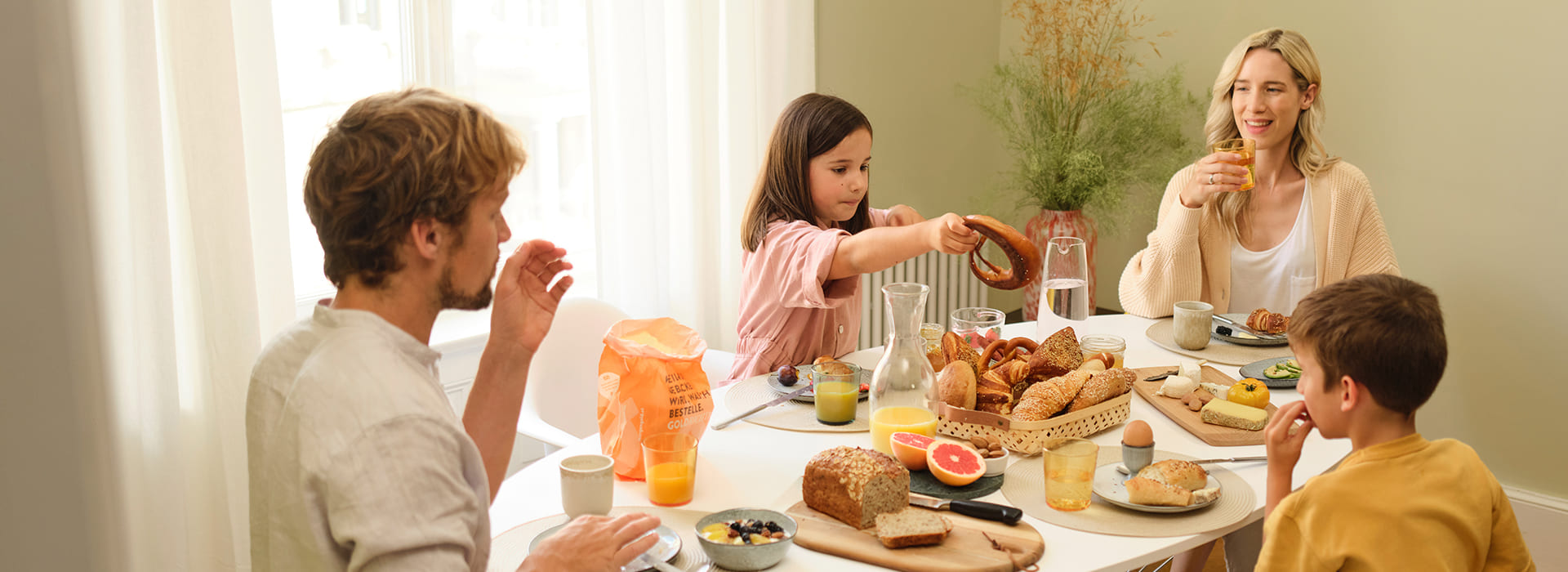 Eine vierköpfige Familie sitzt an einem hellen, gedeckten Frühstückstisch in einem Raum mit grünen Wänden und weißen Vorhängen. Der Tisch ist mit verschiedenen Brotsorten, Gebäck, Obst, Käse, Eiern und Getränken wie Saft und Wasser gedeckt. Ein Mädchen in einem rosa Kleid  greift nach einer Brezel, während ein Junge mit braunen Haaren am Tisch sitzt. Ein Mann mit hellem Haar, im weißen Hemd, sitzt links und isst, während eine blonde Frau im cremefarbenen Cardigan rechts ein Glas Orangensaft hält und lächelt. Im Hintergrund steht eine Pflanze in einer Vase.
