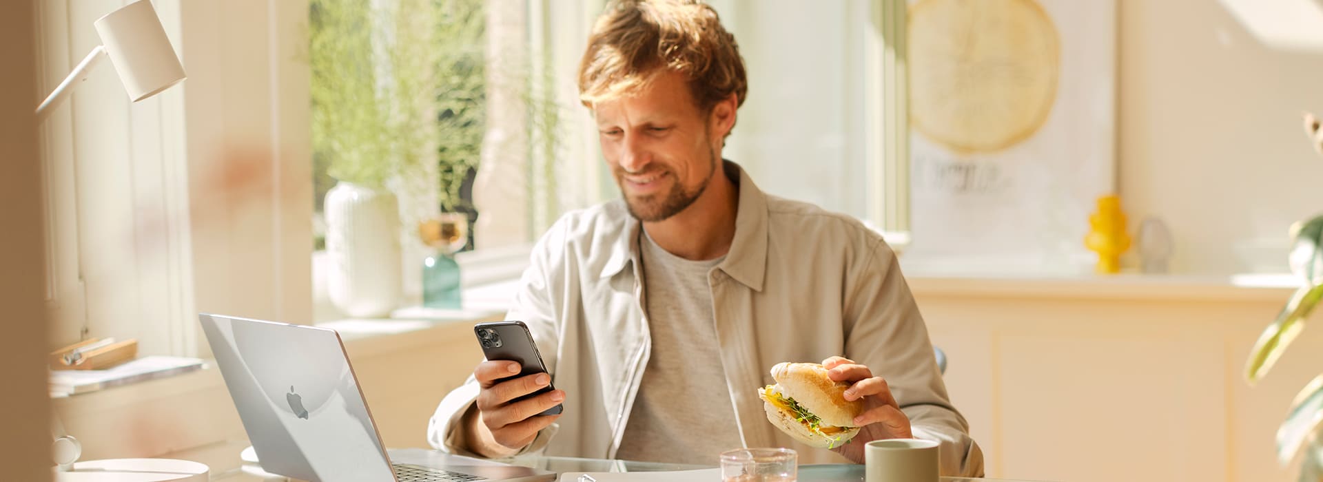 Ein Mann mit mittellangen blonden Haaren und Bart sitzt an einem modernen, lichtdurchfluteten Arbeitsplatz. Er trägt eine helle Jacke über einem grauen T-Shirt und hält in einer Hand ein Sandwich, während er in der anderen Hand ein Smartphone betrachtet und lächelt. Auf dem Glastisch vor ihm stehen ein aufgeklappter Laptop, ein Notizblock, ein Glas Wasser und eine Tasse. Im Hintergrund sind Pflanzen, eine Lampe und ein dekoratives Poster an der Wand zu sehen.