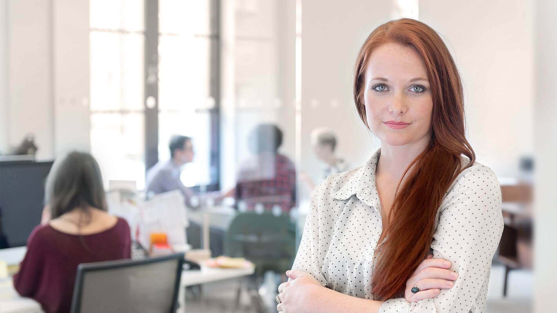 Frau mit langen roten Haaren steht selbstbewusst mit verschränkten Armen in einem modernen, hellen Büro und blickt direkt in die Kamera. Sie trägt eine weiße Bluse mit kleinen Punkten. Im Hintergrund sind unscharf weitere Personen an Schreibtischen zu sehen, die gemeinsam arbeiten.