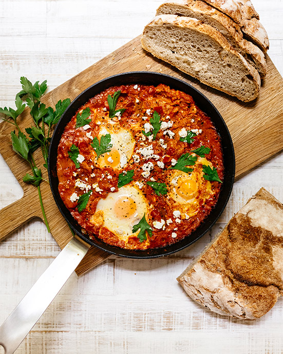 Pfanne mit Shakshuka Tomatensauce Ei Petersilie und Brot auf Holzbrett.