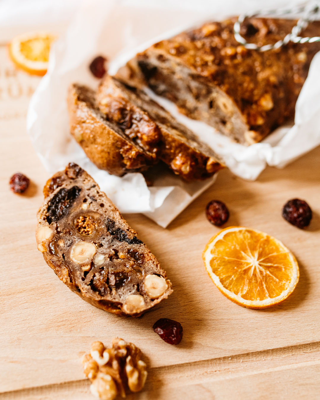 Anschnitt eines fruchtigen Brotes mit Nüssen, getrockneten Feigen, Datteln und Haselnüssen auf einem Holzbrett, dekoriert mit Trockenfrüchten, Walnüssen und einer getrockneten Orangenscheibe.