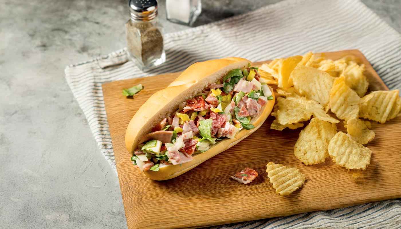 Belegtes Sandwich mit Salami, Schinken, Käse und Salat, serviert mit Pommes frites auf Holzbrett.