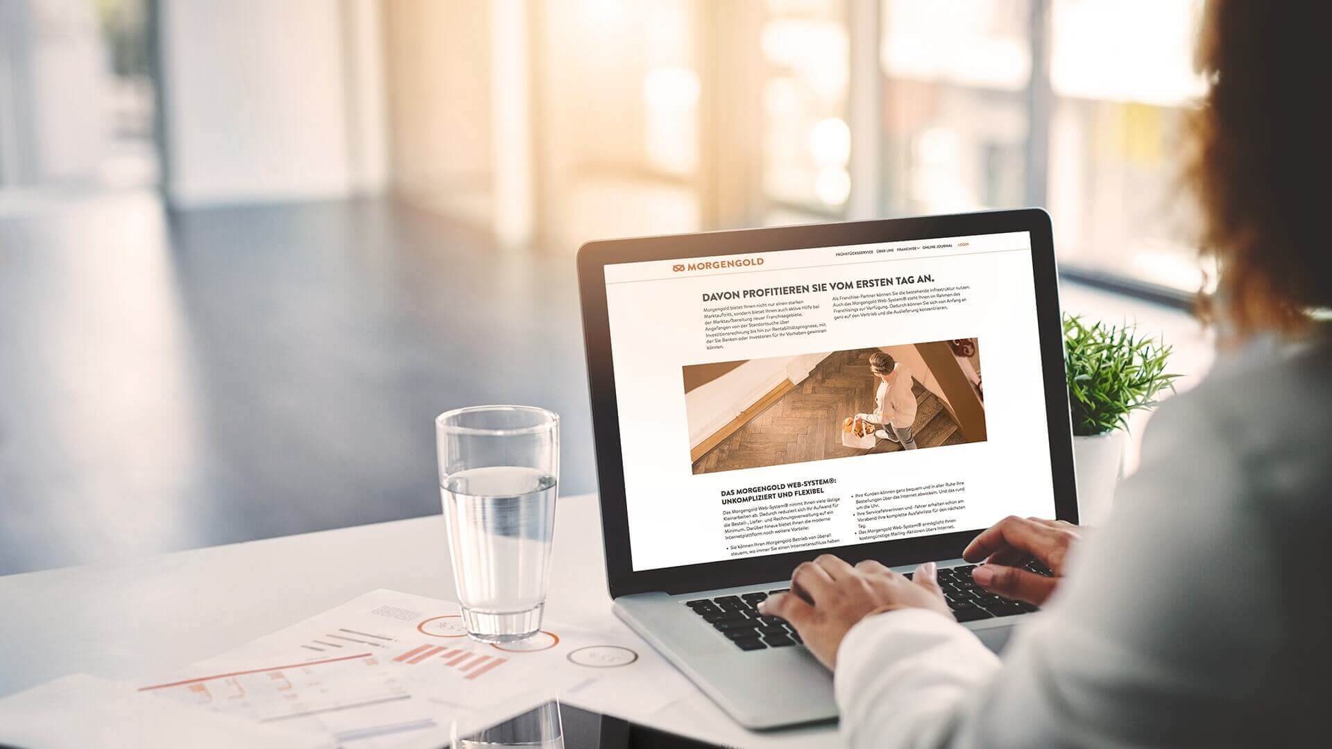 Morgengold - Über Uns Person sitzt in einem hellen Raum mit großer Fensterfront an einem Schreibtisch vor einem geöffneten Laptop. Auf dem Bildschirm ist eine Franchise-Informationsseite von Morgengold zu sehen.